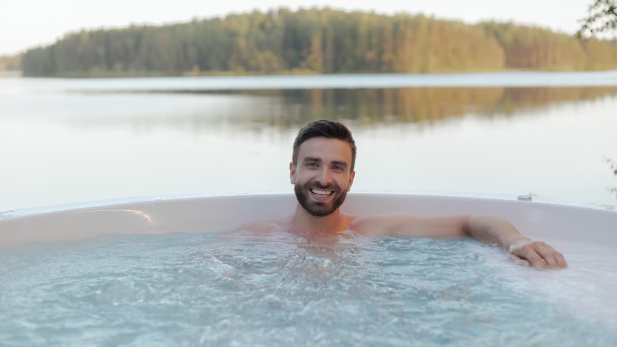 Shirtless man smiling in an outdoor hot tub by a serene lake, enjoying relaxation and wellness.