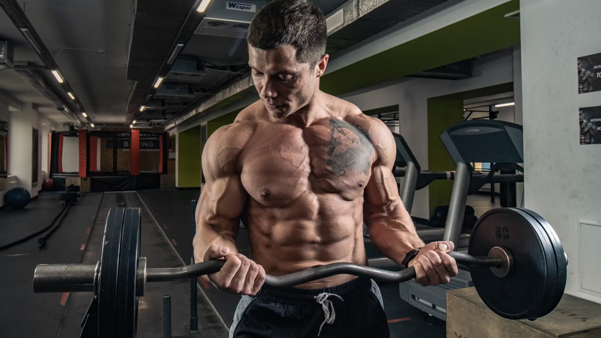 Topless bodybuilder with tattoos lifting weights in an indoor gym setting.